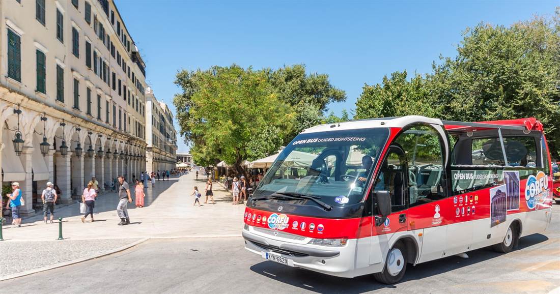 A small red and white open-top tour bus on a paved street beside an arcaded building and trees in Corfu Town, part of Corfu hop on hop off.