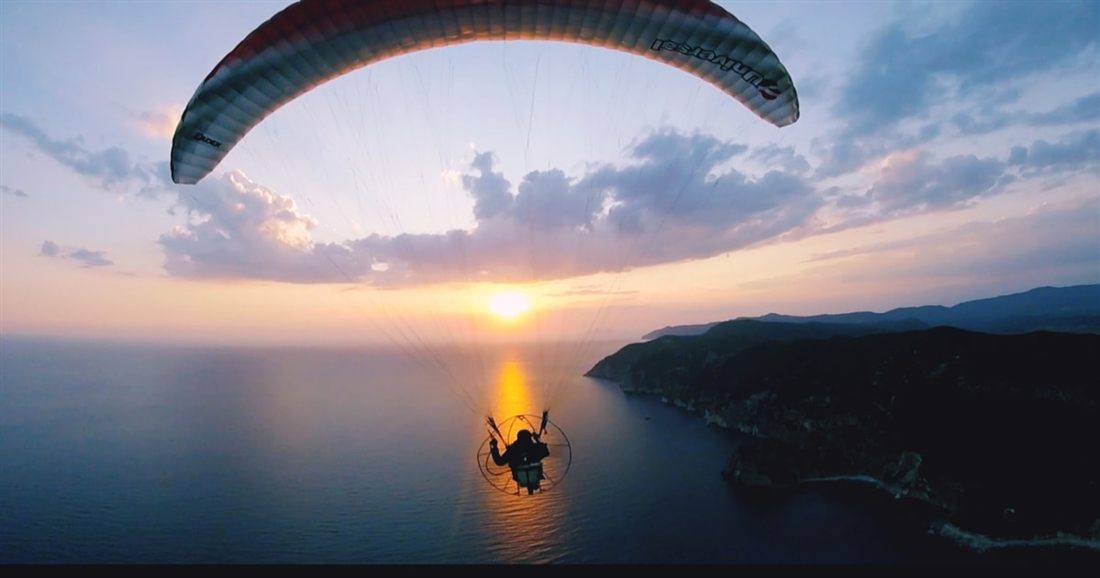 A paratrike flies under its wing over the sea at sunset, with the sun low on the horizon and a dark headland to the right, on Corfu paratriking.