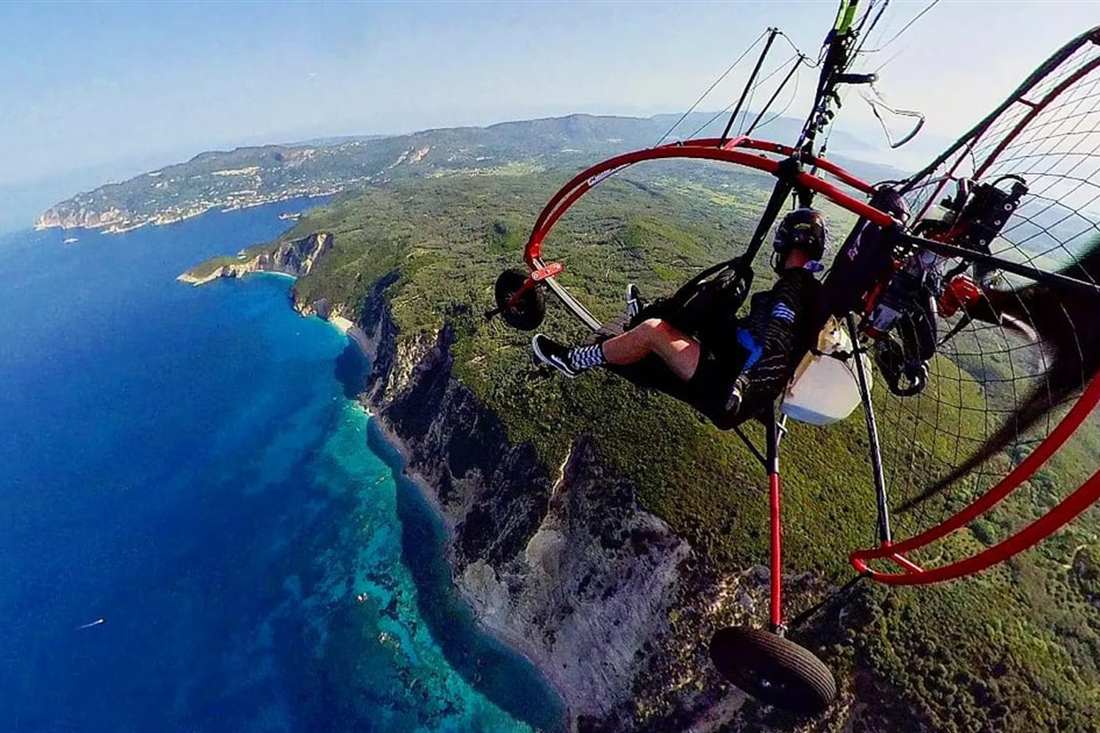 A pilot and passenger fly in a red paratrike above high green cliffs and deep blue sea on the west coast during Corfu paratriking.