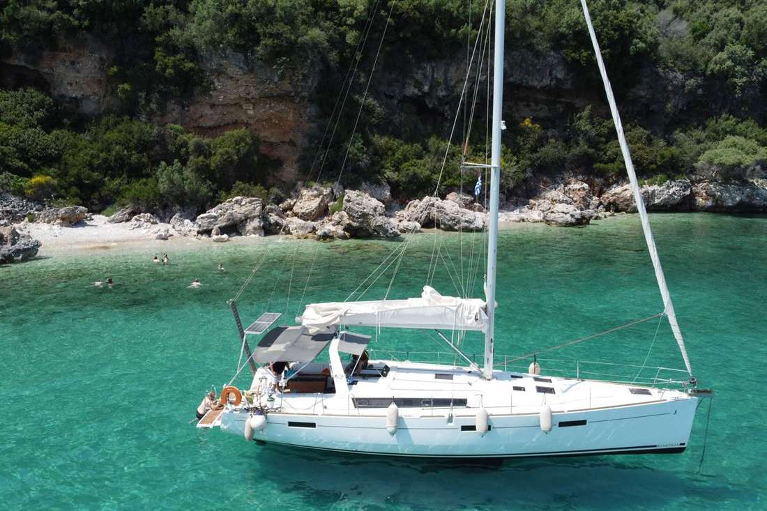 A white sailing yacht anchored in a turquoise cove below a wooded rocky shore, with swimmers near a small beach