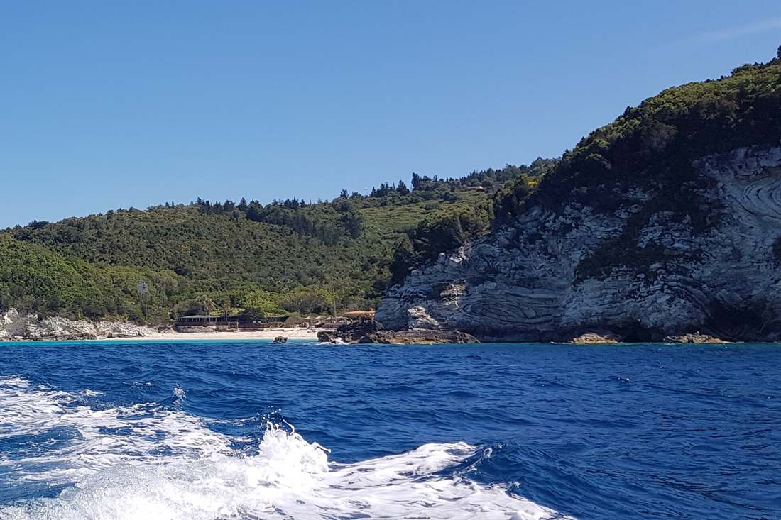 White rock cliffs and a green wooded headland with a sandy beach seen from a boat, with the wake in the deep blue sea