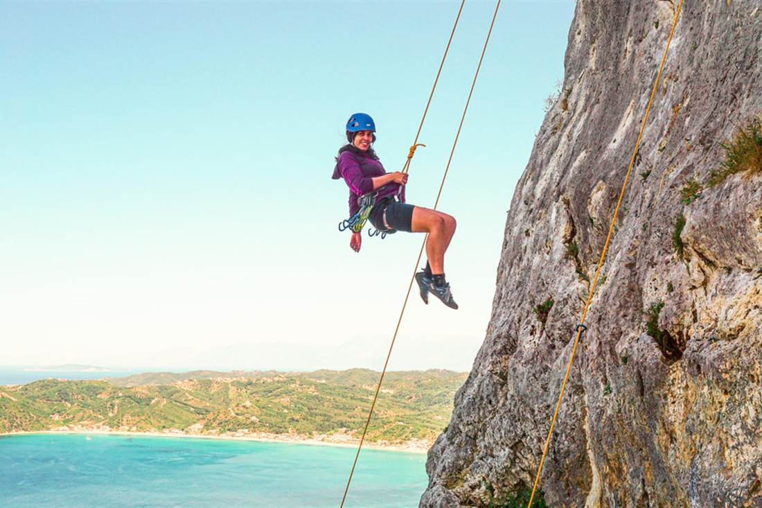 A climber in a helmet abseiling down a limestone cliff high above a bay with clear blue water on Corfu rock climbing.