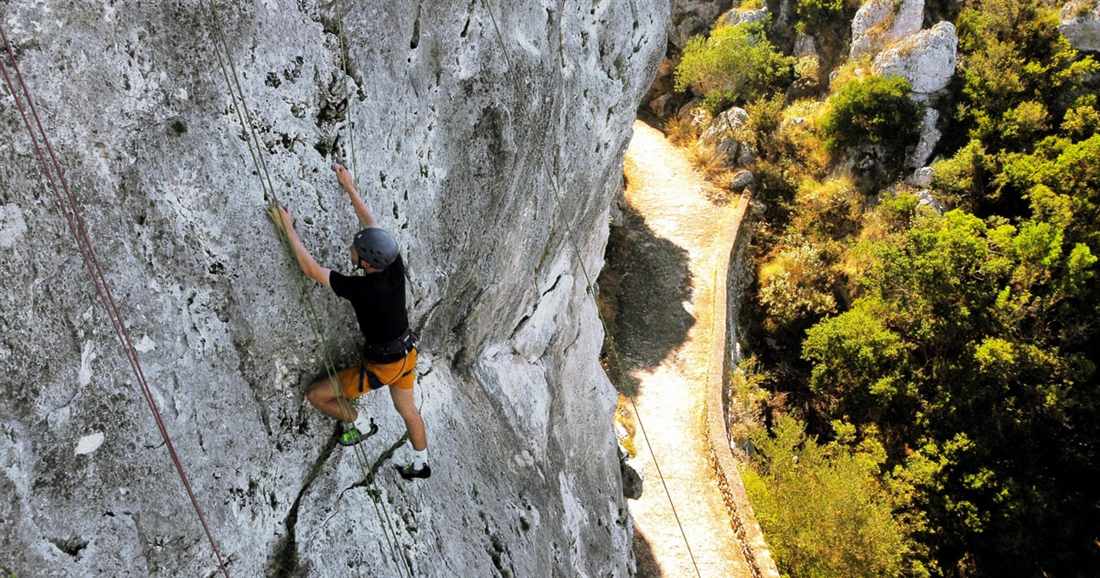 A climber in a helmet moving up a grey limestone wall above a path near Paleokastritsa, on a Corfu rock climbing session.