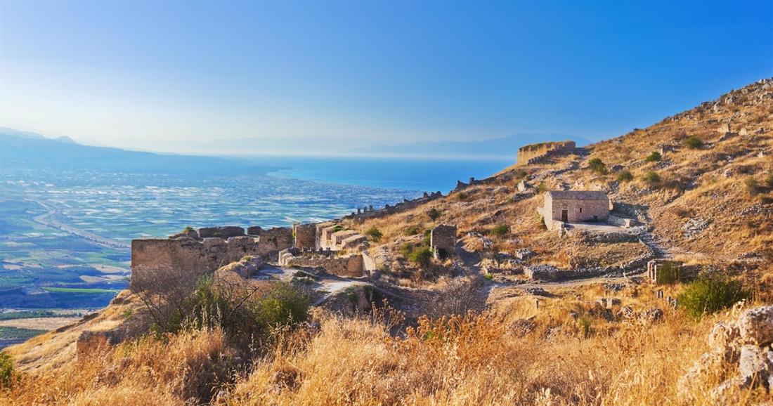 Stone walls and towers of the Acrocorinth fortress on a dry hillside, with the plain and the Gulf of Corinth below.