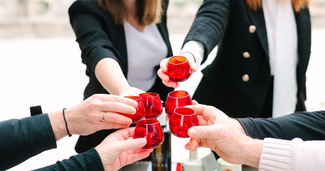 Hands raise small red glass tasting cups together over a table, at a Corinth olive oil tasting.