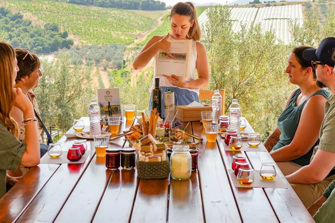 A host reads from a card at a long wooden table set with red tasting cups, bottles and jars, as guests sit under olive trees above green hills, at a Corinth olive oil tasting.
