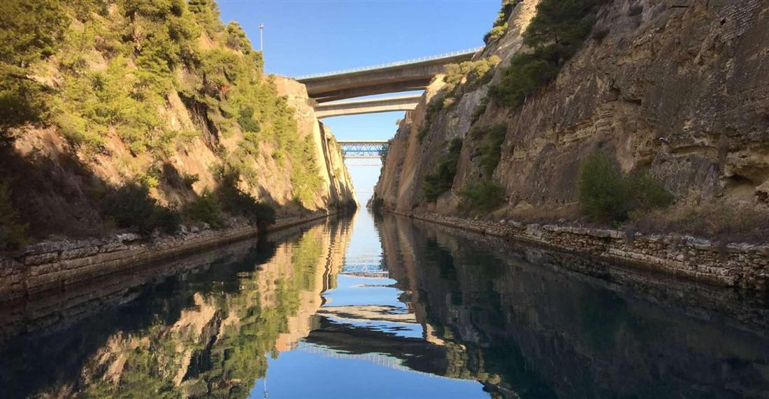 The still water of the Corinth Canal between its high rock walls under a road bridge