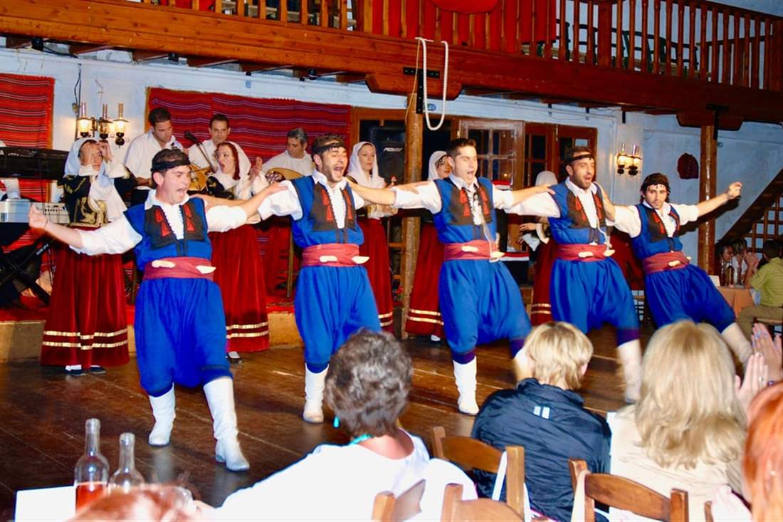 Dancers in red, blue and white Cretan costume performing a line dance on stage before diners at a Cretan night.