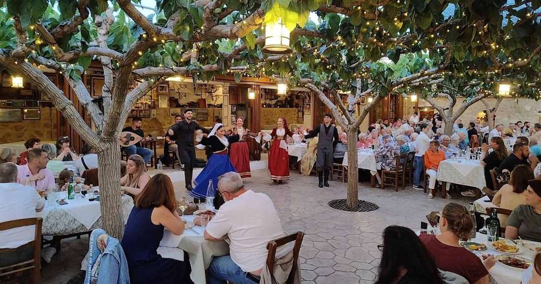 Guests at dinner tables under the trees of a village courtyard while dancers in Cretan costume perform, at a Cretan night.