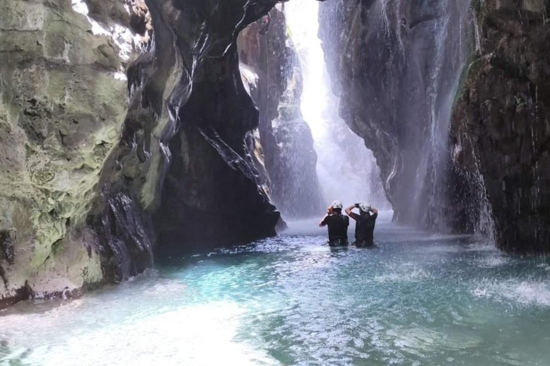 Two canyoners wading through a turquoise pool below a waterfall in a narrow rock gorge on a Crete canyoning tour.