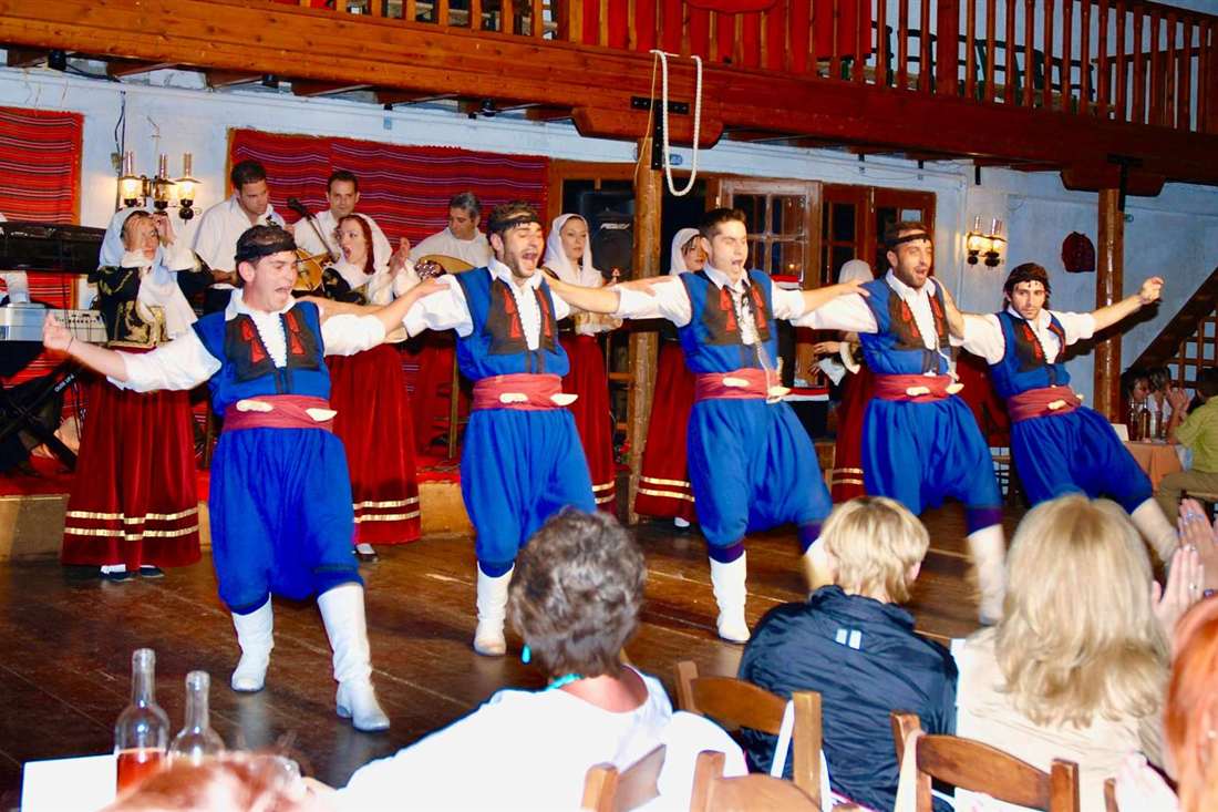 Five dancers in blue vests, red sashes and white boots dance a line dance on a wooden stage in Crete while musicians play behind them