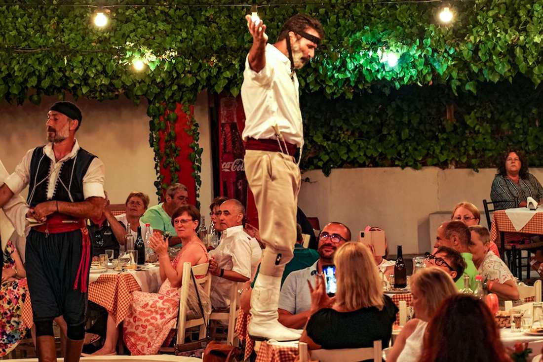 A Cretan dancer in white shirt and boots stands raised above the tables while diners clap and film under vine-covered lights at a taverna evening