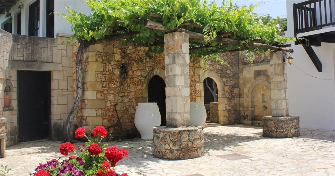 A stone village courtyard with a vine-covered pergola, a stone well, clay jars and red geraniums, on a Crete private tour.