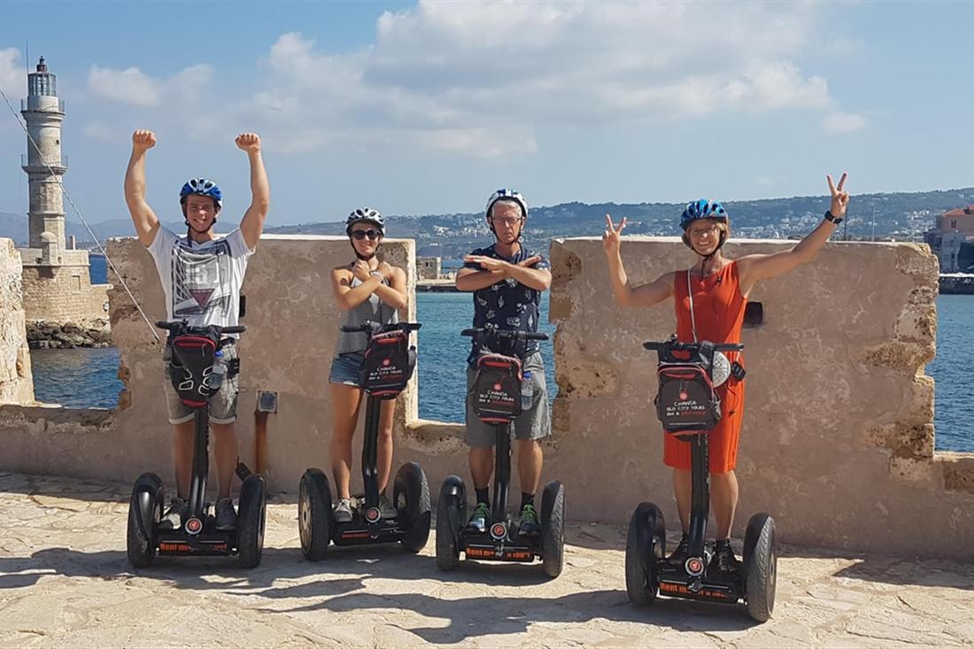 Four riders in helmets on segways on the harbour wall in Chania, with the Venetian lighthouse behind, on a Crete segway tour.