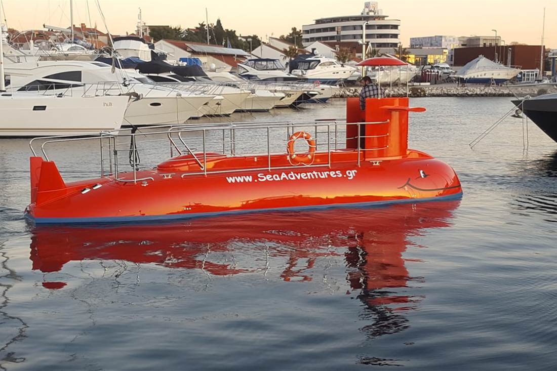 A red semi-submarine boat with portholes floats in a harbour beside moored yachts, used for a Crete submarine trip.