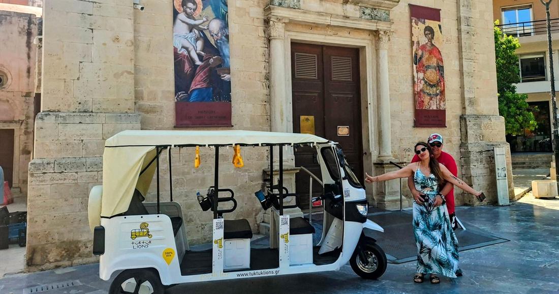 An electric tuk tuk parked in front of a stone church facade, with two visitors beside it, on a Heraklion tuk tuk city tour.