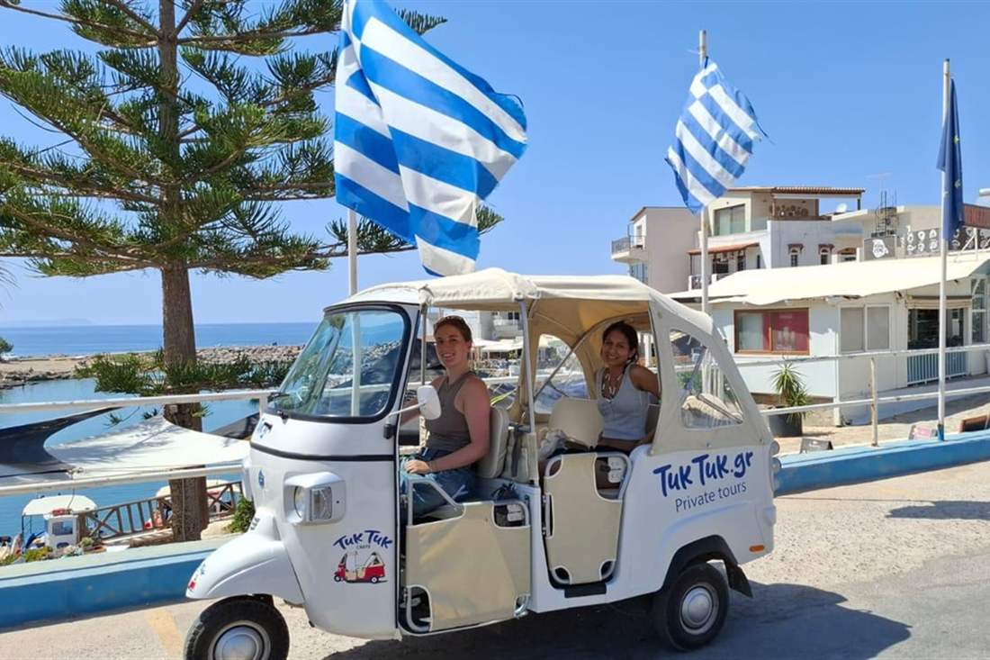 A white tuk tuk with two passengers parked beside Greek flags and the sea on a Crete tuk tuk tour.
