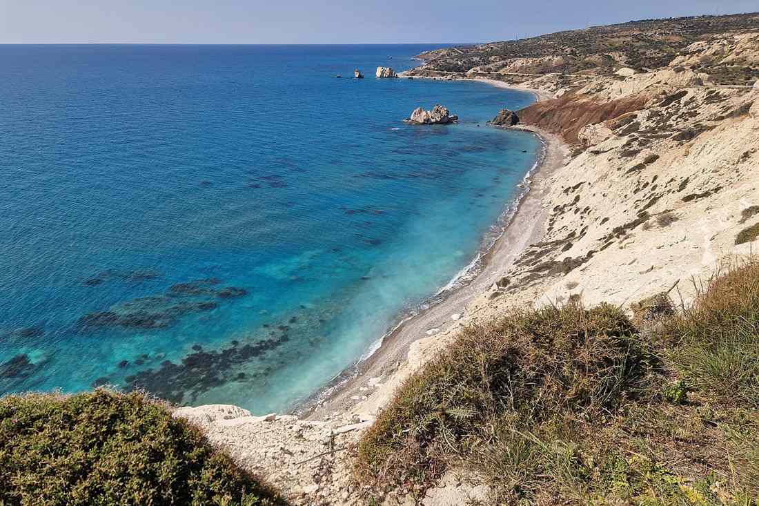 Blue sea, a pebble beach and white rocky cliffs with sea stacks offshore and a coast road on the headland