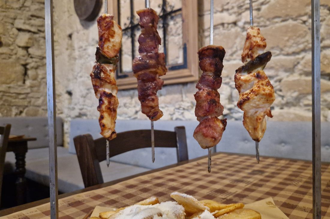 Four chicken and pork souvlaki skewers hanging on a stand above a checked tablecloth in a stone-walled taverna, with chips in front