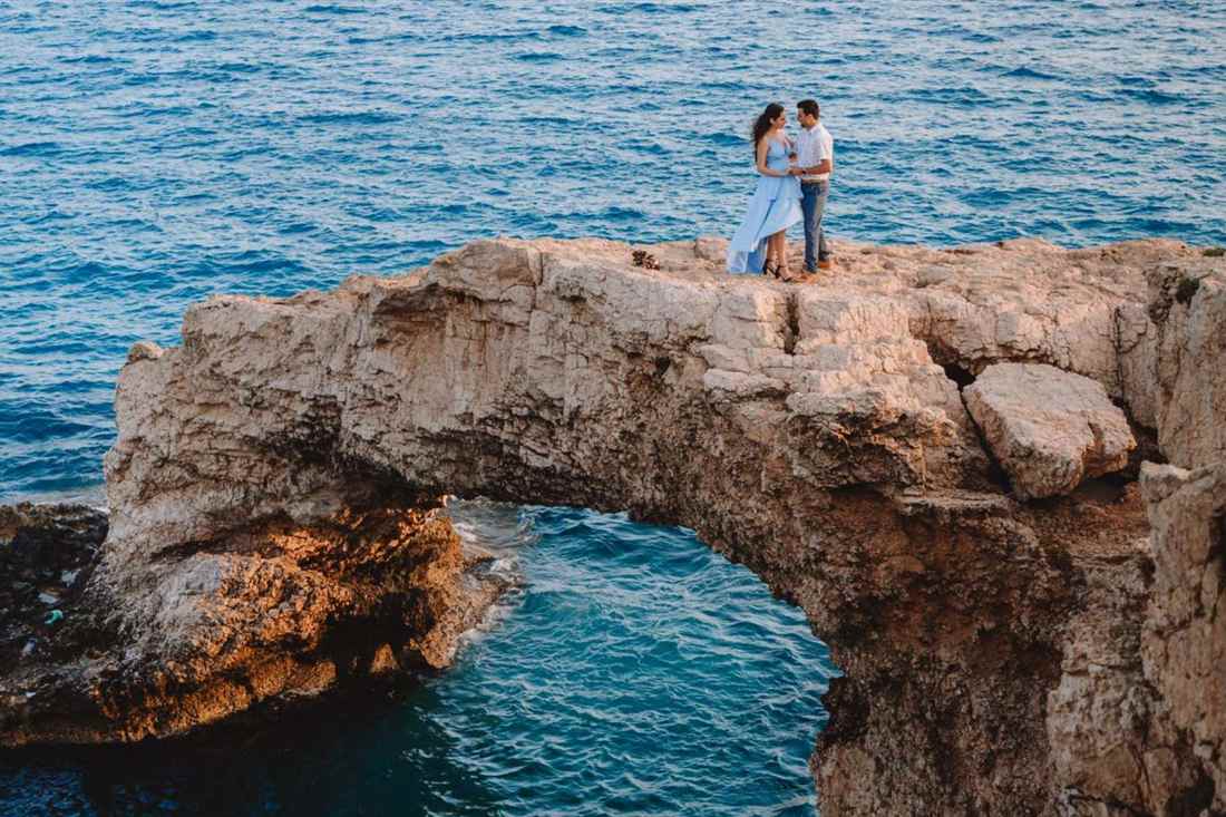 A couple standing close together on top of a natural rock arch above deep blue sea, the woman in a light blue dress and the man in a white shirt and jeans