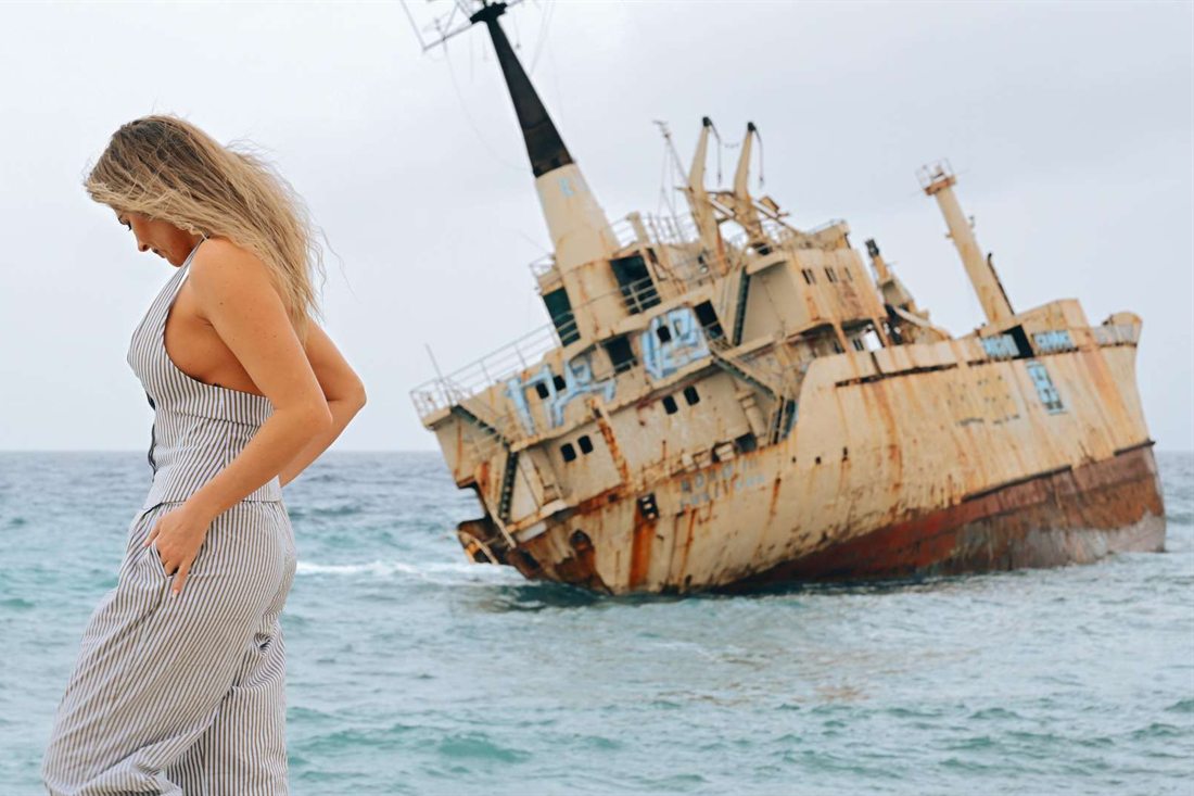 A woman in a striped outfit looking down on the shoreline, with a rusted shipwreck tilted in the turquoise sea behind her under a grey sky