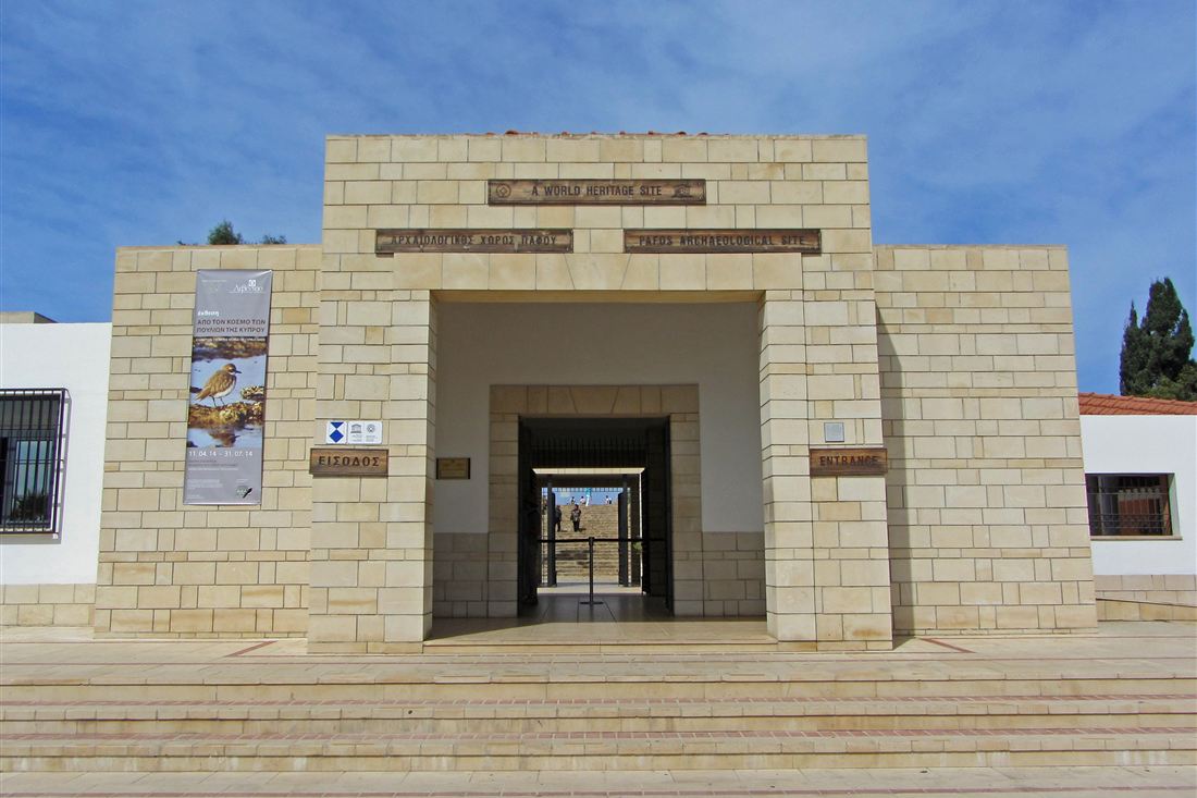 Stone entrance building of the Pafos Archaeological Site with a World Heritage Site sign, an Entrance sign and wide steps under a blue sky