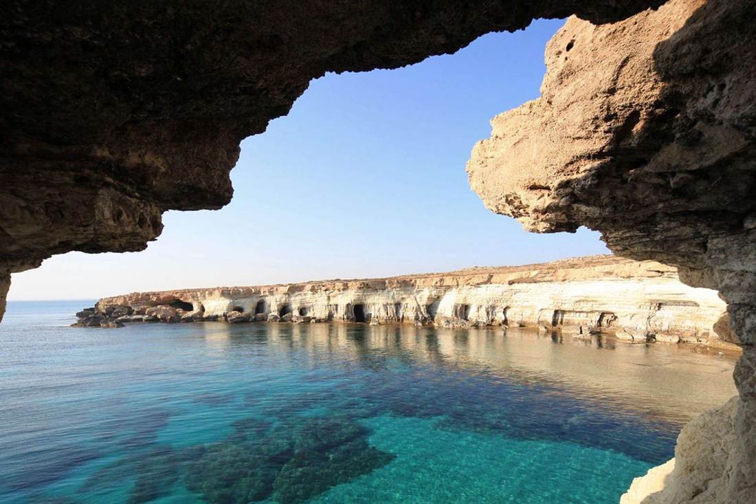 Sea caves in the limestone cliffs of Cape Greco seen through a rock arch above clear turquoise water