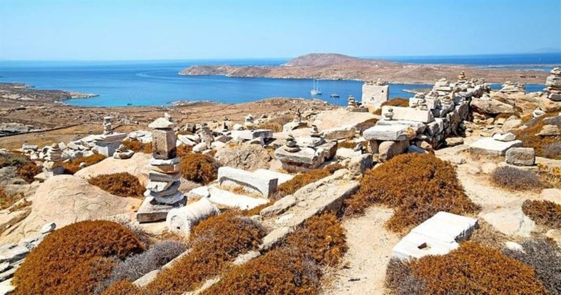 Low stone ruins and scattered marble blocks cover a dry hillside on Delos, with the blue sea and a neighbouring island behind.