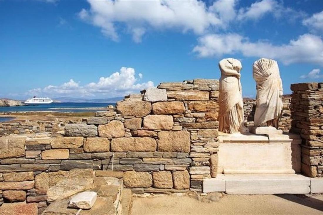 Two headless marble statues stand on a stone base among ruined walls on Delos, with the sea and a white ship behind.