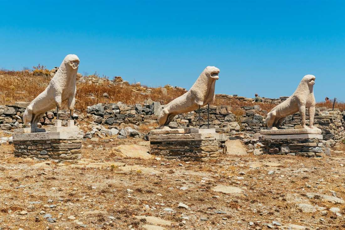 Three marble lions sit in a row on a dry stone terrace under a blue sky on Delos.