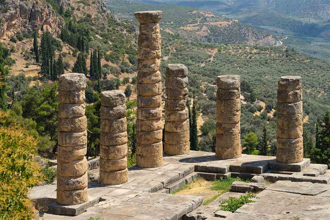 Six standing column drums of the Temple of Apollo at Delphi on their stone platform, with cypress trees and the Pleistos valley below.