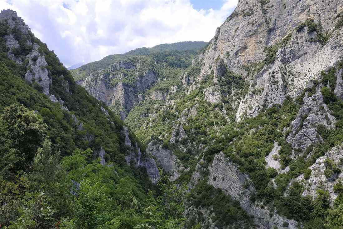 A deep green limestone gorge of the Enipeas river on Mount Olympus, with steep grey rock walls under a bright cloudy sky