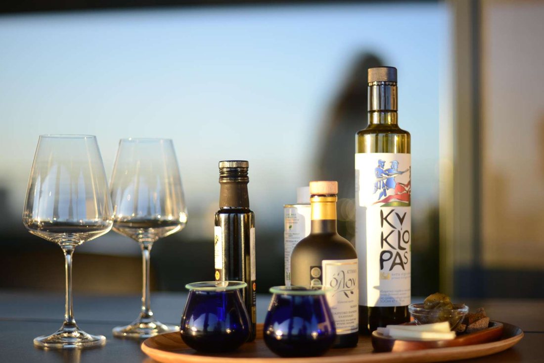 Two blue olive oil tasting glasses, bottles of Greek olive oil and two wine glasses on a wooden tray in evening sunlight