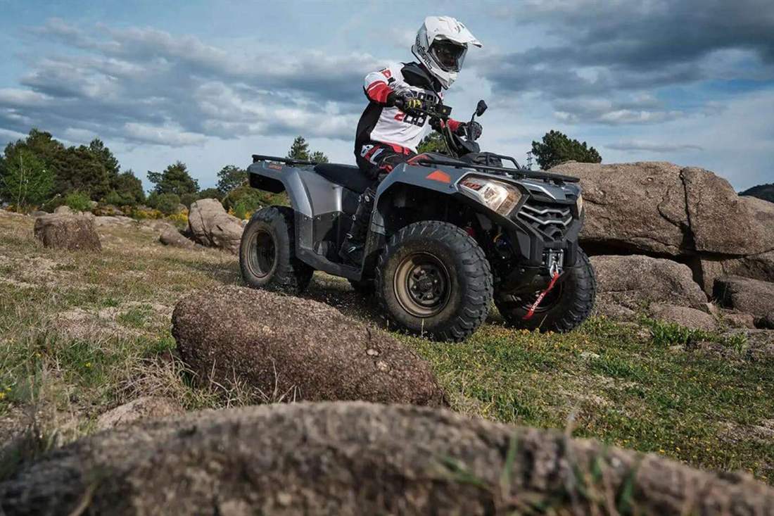 A rider in a white helmet and racing suit drives a grey ATV between large rounded boulders on a grassy slope under a cloudy sky