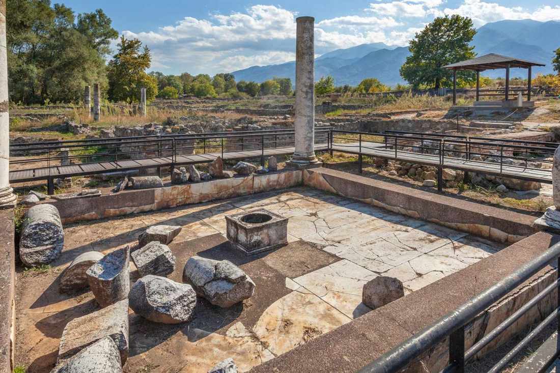 Marble floor, fallen column drums and a standing column at the Dion archaeological site, with walkways and mountains behind
