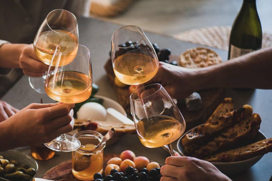 Four hands clink glasses of white wine over a table with cheese, grapes, olives, apricots and bread