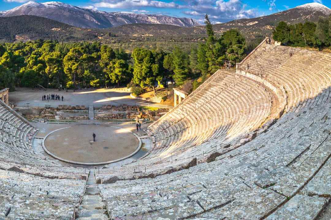 The ancient theatre of Epidaurus seen from the upper rows, with the round orchestra, pine trees and snow-capped mountains behind.