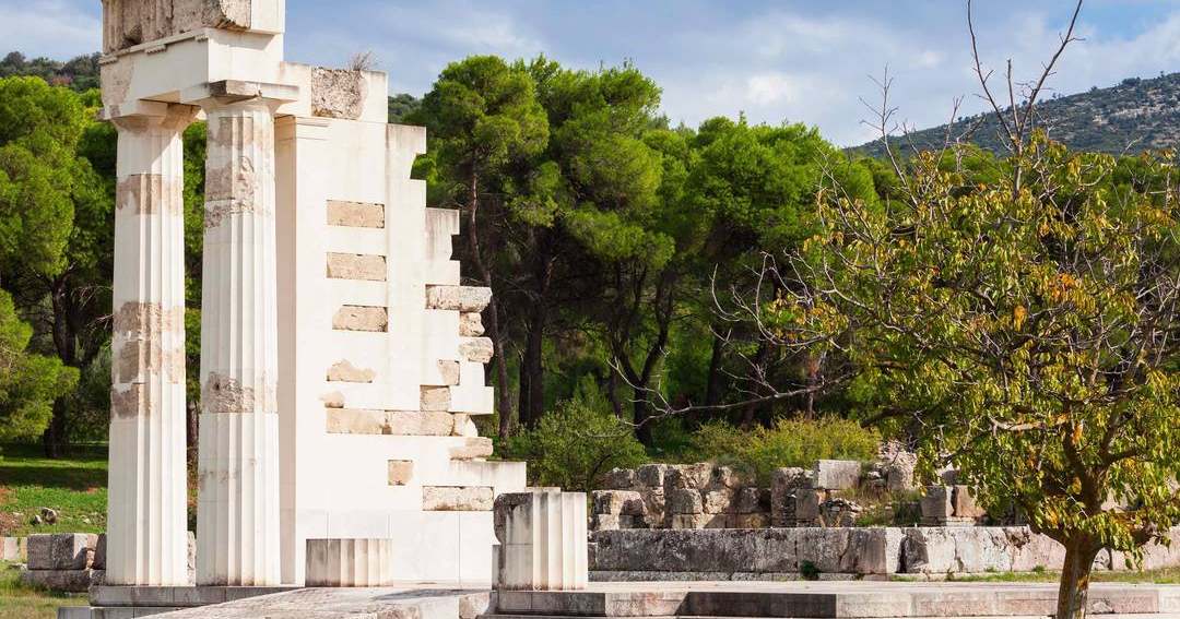 Restored Doric columns and a stepped stone wall in the Sanctuary of Asclepius at Epidaurus, with pine trees behind.