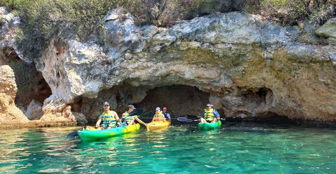 Kayakers in yellow and green sea kayaks paddling by a rock sea cave on turquoise water near Epidavros