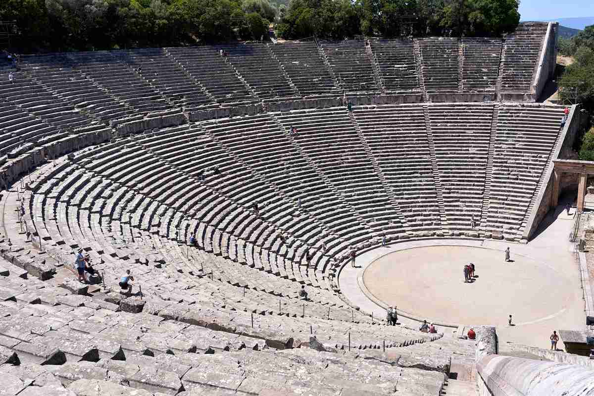 Visitors walk the round orchestra below the curved stone seats of the ancient theatre of Epidaurus, the monument at the heart of every Epidaurus tour.