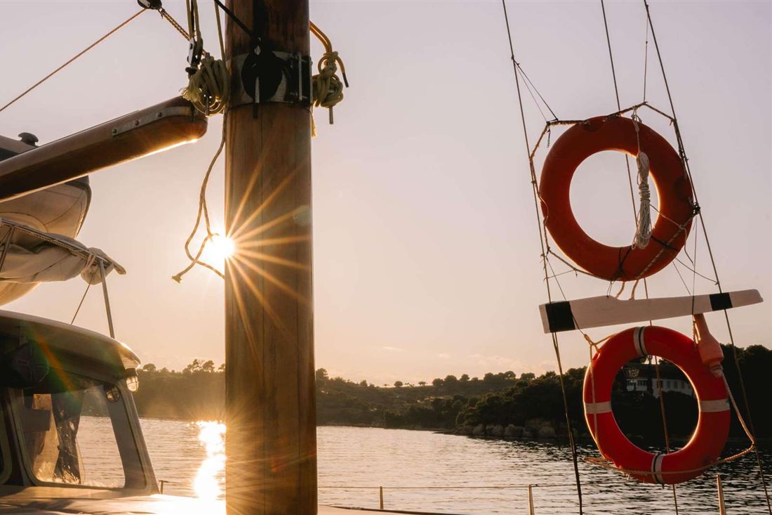 Sun setting behind the wooden mast of a sailing boat in a pine-lined cove, with orange lifebuoys hanging from the rigging
