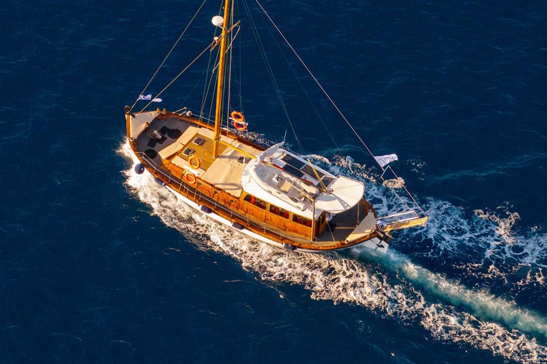 Wooden sailing boat cruising on deep blue water in warm evening light, seen from above with a white wake behind it