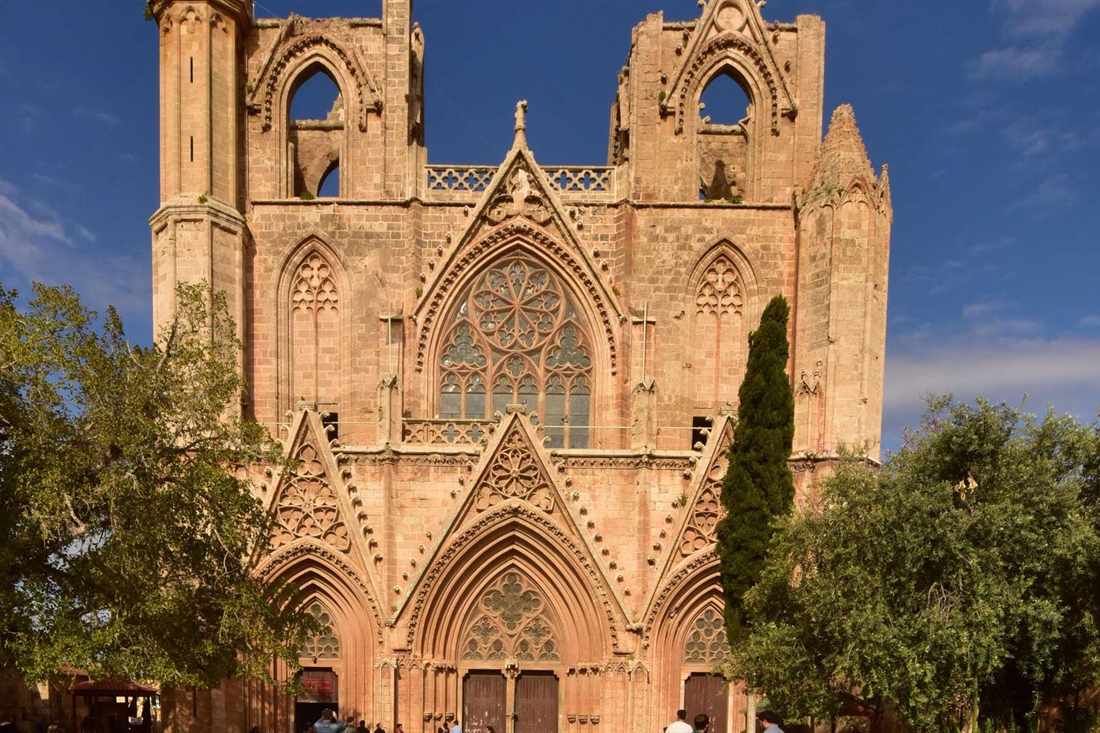 Gothic west facade of the Lala Mustafa Pasha Mosque in Famagusta with two towers, a rose window and three pointed doorways under a blue sky, olive trees on both sides