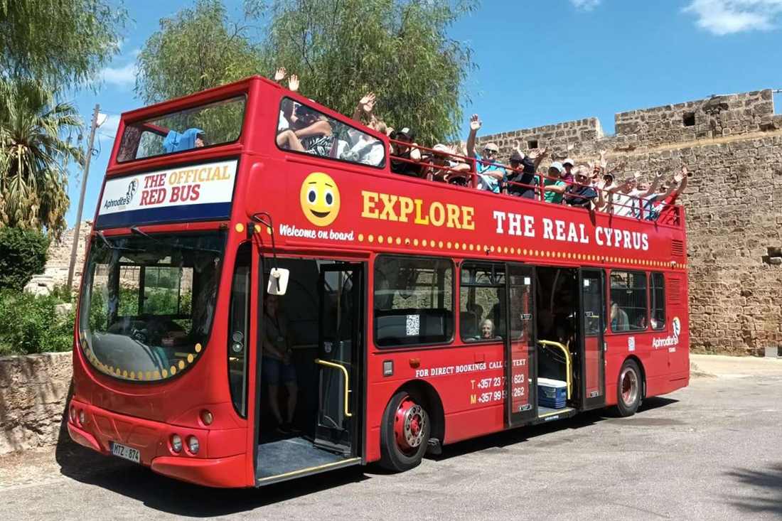 Red open-top double-decker tour bus parked beside the old stone walls of Famagusta, with passengers waving from the upper deck