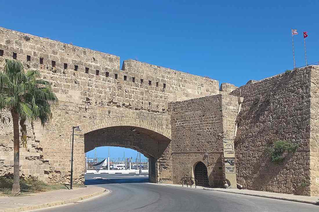 Sandstone Venetian wall of Famagusta with an arched gate, a palm tree and flags under a clear blue sky