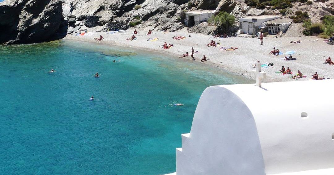 Swimmers in clear turquoise water off a pebble beach on Folegandros, beside a whitewashed chapel.