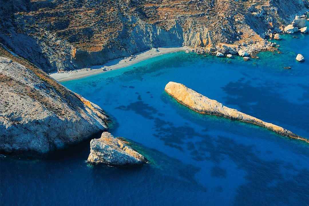 The deep blue bay of Katergo on Folegandros, with a long pale rock in the water below steep cliffs.