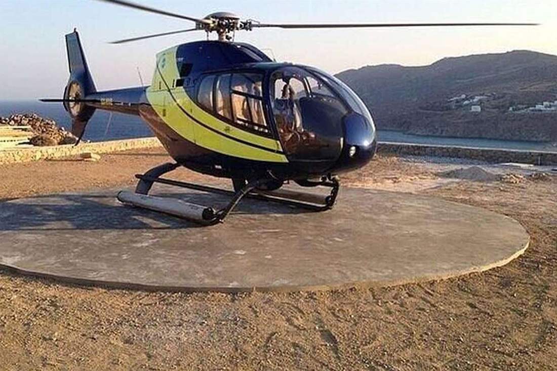 Blue and yellow helicopter parked on a round concrete helipad above the sea, with a hillside and white houses behind it in golden evening light