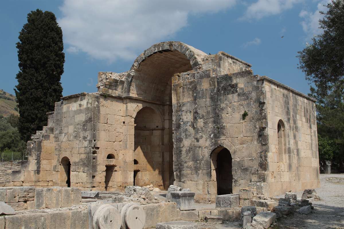 The arched stone apse of the Basilica of Agios Titos rises beside a tall cypress under a blue sky at the archaeological site that Gortyna tickets open to visitors.