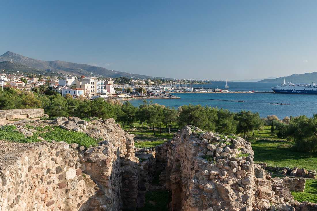 Ancient stone walls in the foreground above Aegina harbour, with pine trees, white houses, a marina, a white ferry on blue water, and mountains behind